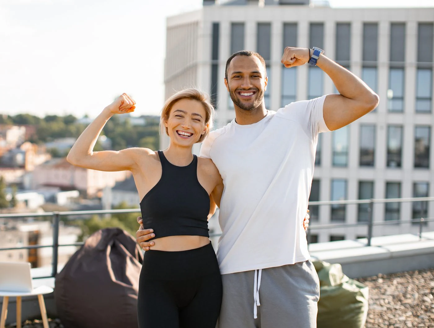 A smiling couple flexing their arms together on a rooftop, celebrating their fitness progress