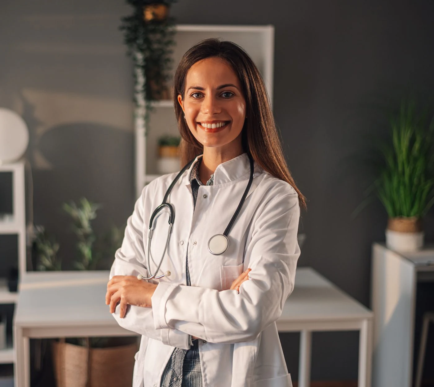 Confident female physician in a white coat with a stethoscope, smiling warmly with arms crossed in a calm clinical office