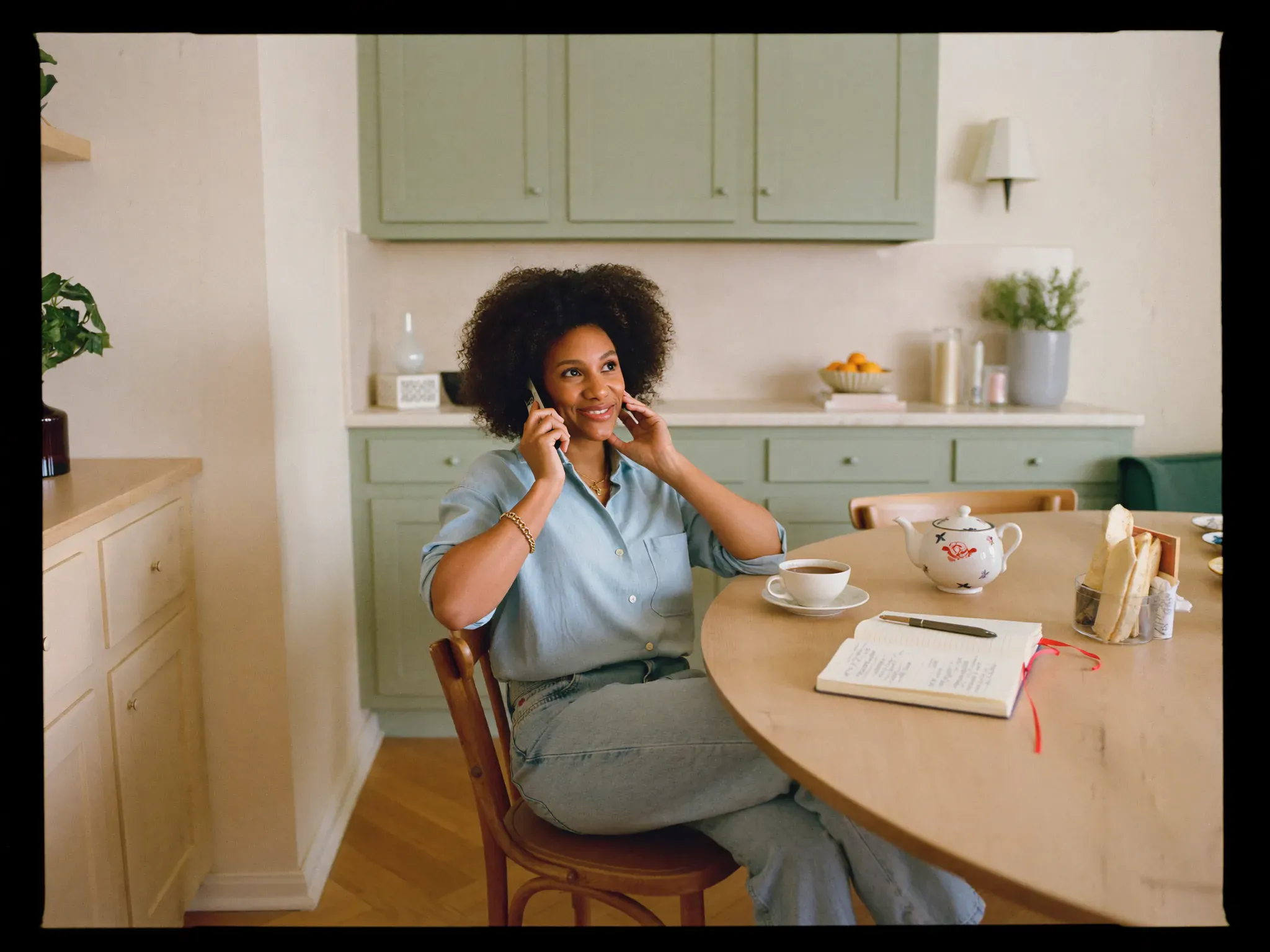 A relaxed adult chatting warmly on a phone call at a sunny kitchen table with tea and a notebook nearby